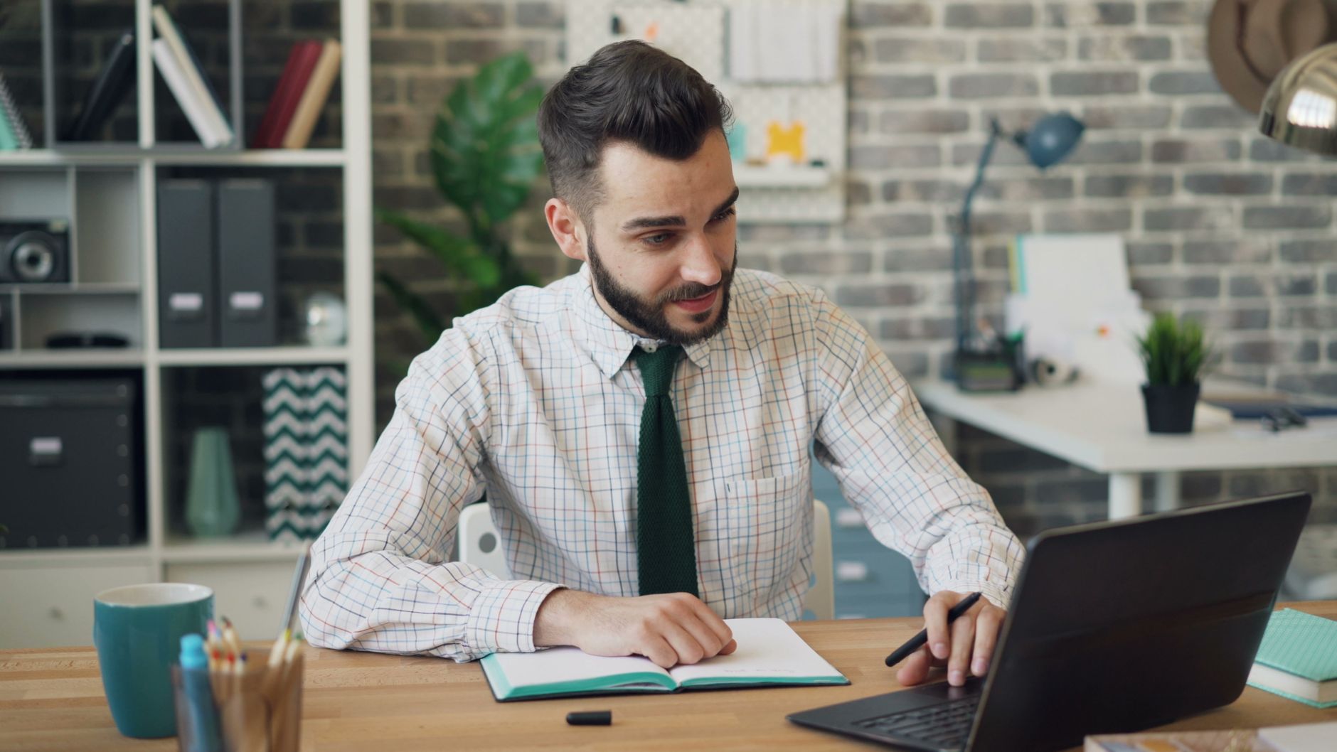 A bearded man in a checked shirt and green tie smiles at his laptop, an open notebook and pen on a wooden desk in a brick-walled office.
