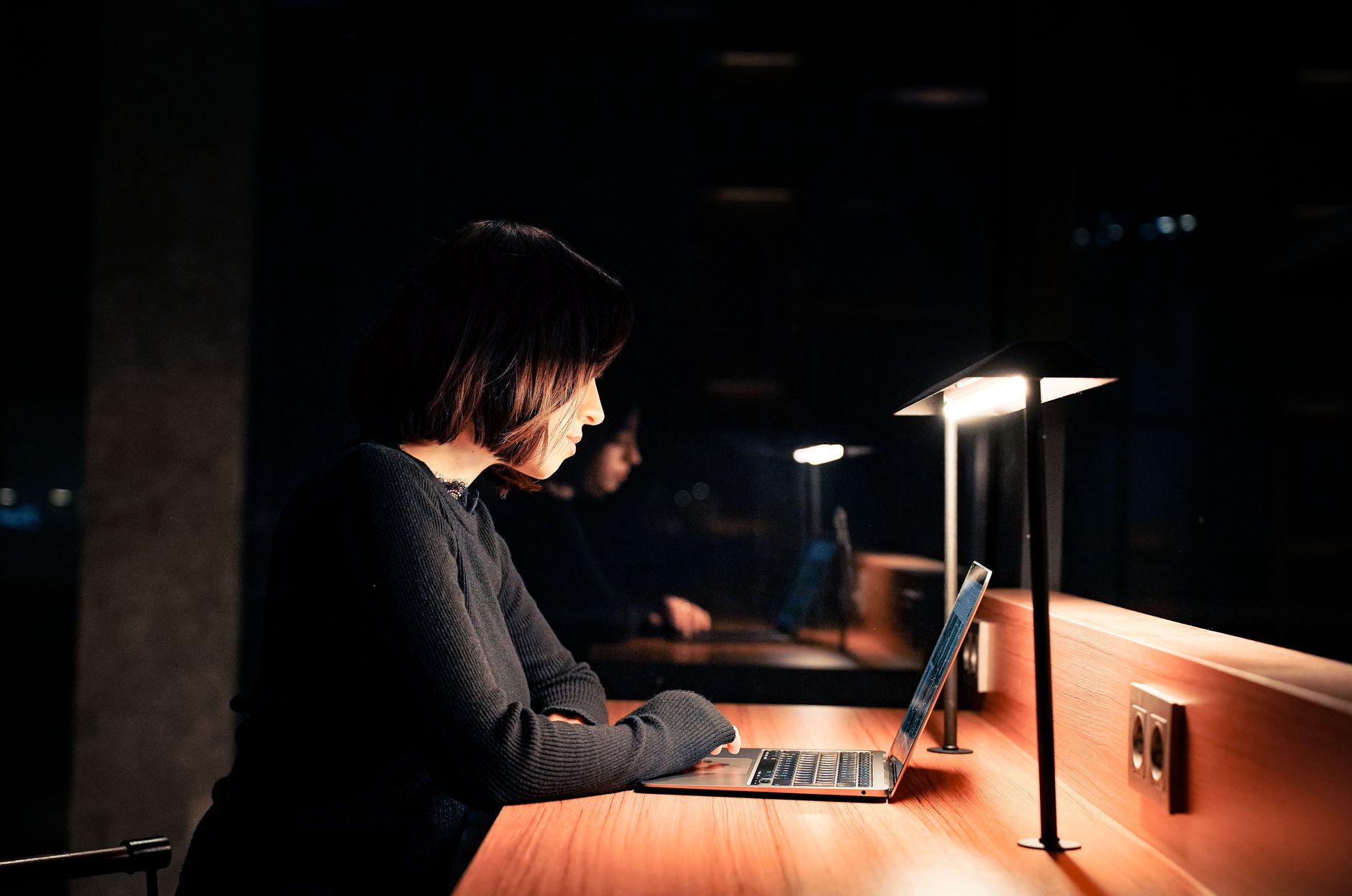 A woman with a dark bob sits side-on at a wooden desk in a dark room, her laptop and a desk lamp lit warmly.