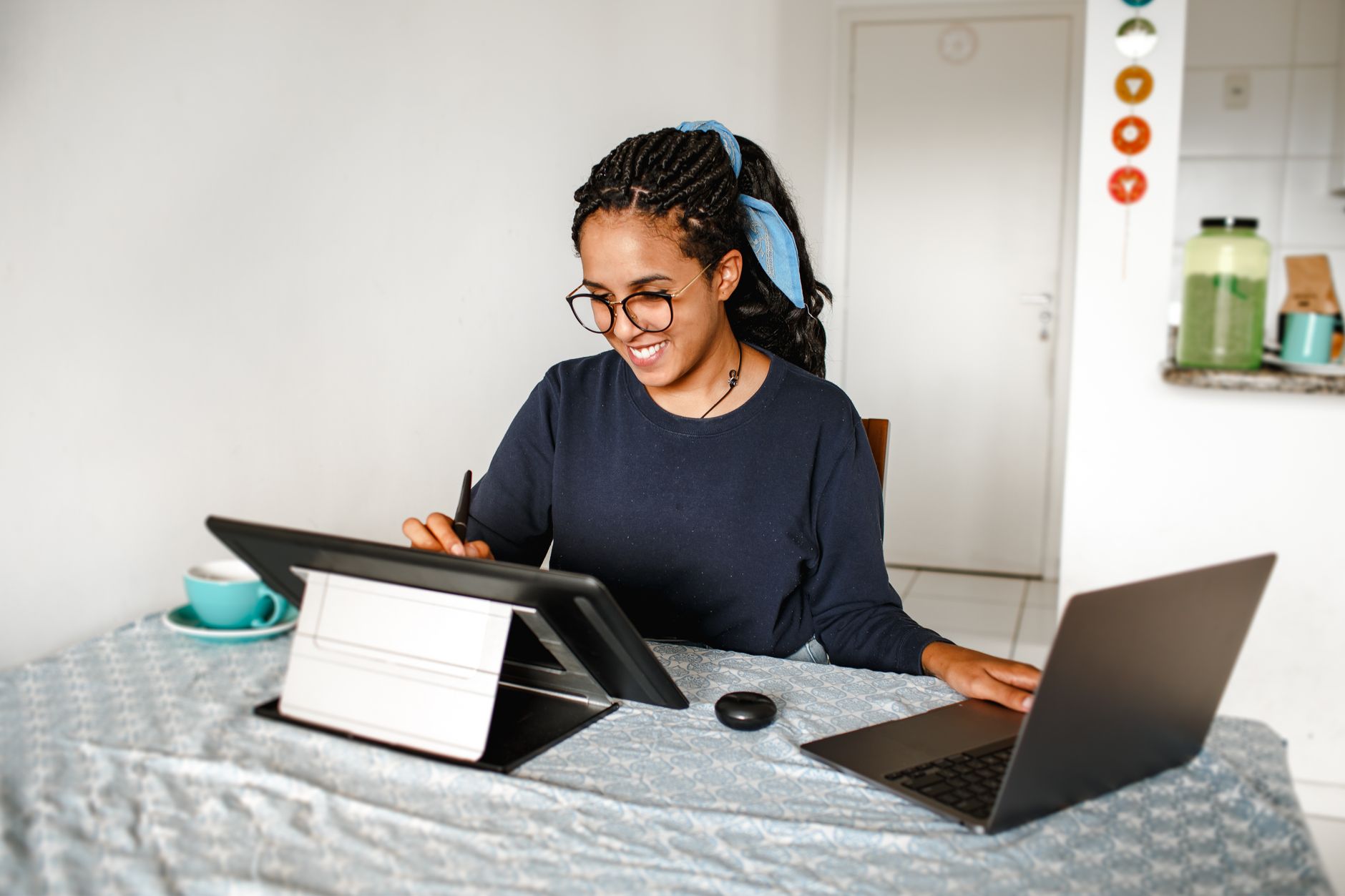 A smiling young woman in glasses and a blue hair scarf works at a laptop and a drawing tablet at a table in her home.