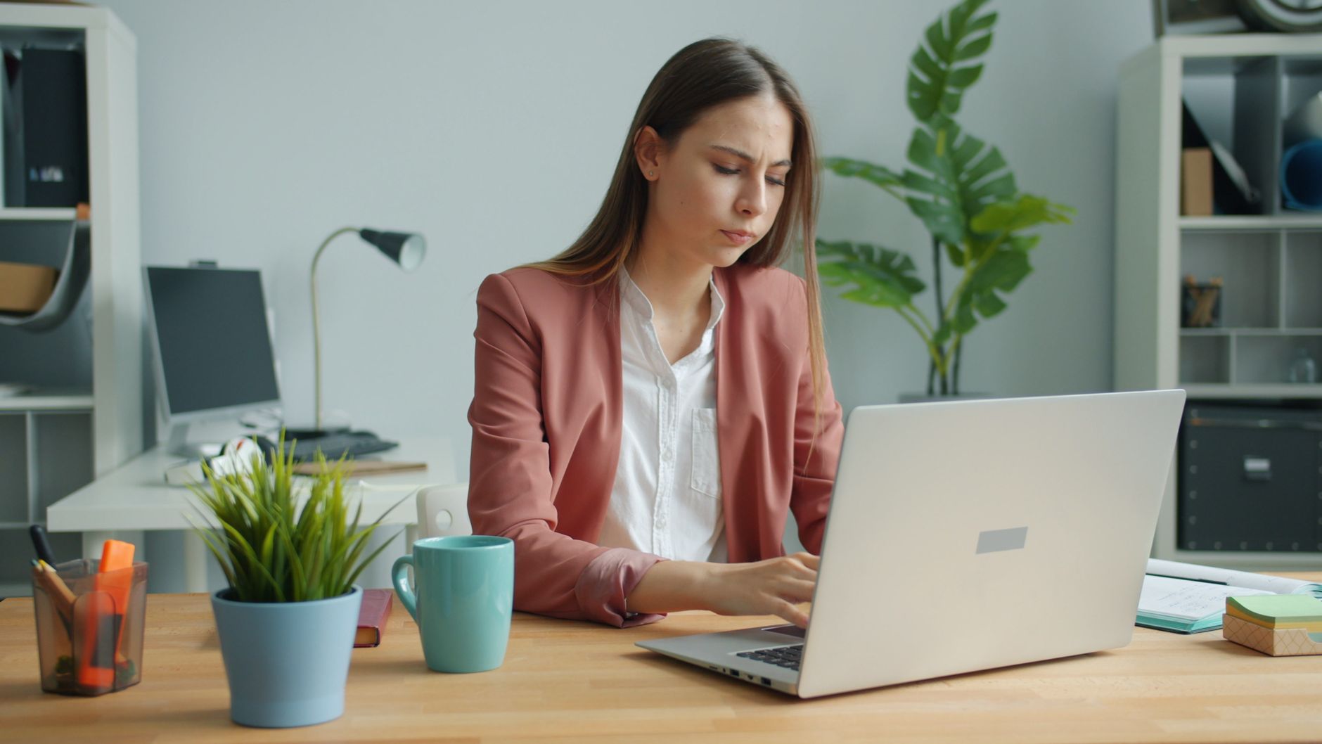 A young woman in a pink blazer concentrates on her laptop at a wooden desk with a teal mug, a plant and a lamp.