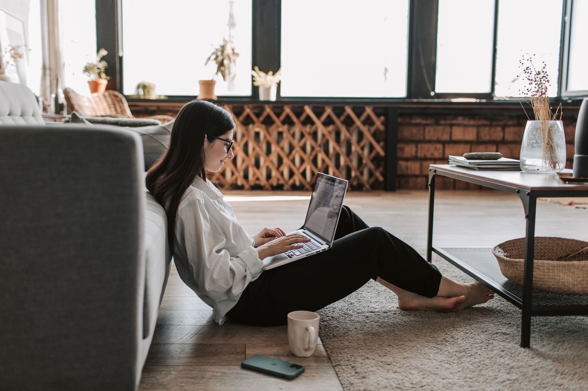A young woman with long dark hair sits on the living-room floor with a laptop on her knees, a mug and a phone beside her.