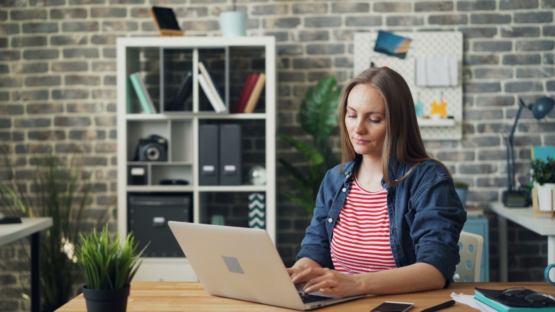 A woman in a denim jacket and red-striped top types on a silver laptop at a desk in front of a brick wall and a white shelving unit.